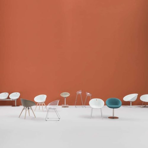 Serie di sedie della stessa famiglia disposte orizzontalmente su un piano bianco con sfondo arancione e azzurro